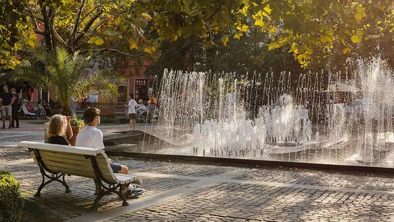 People sitting on a bench in a public square with a water feature shooting water into the air.