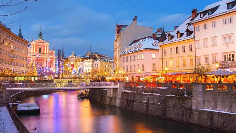 Buildings along the river in Ljubljana with snow on their rooves and Christmas lights up.