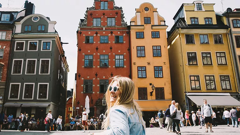 Person wearing sunglasses stands in front of a row of brightly coloured buildings.