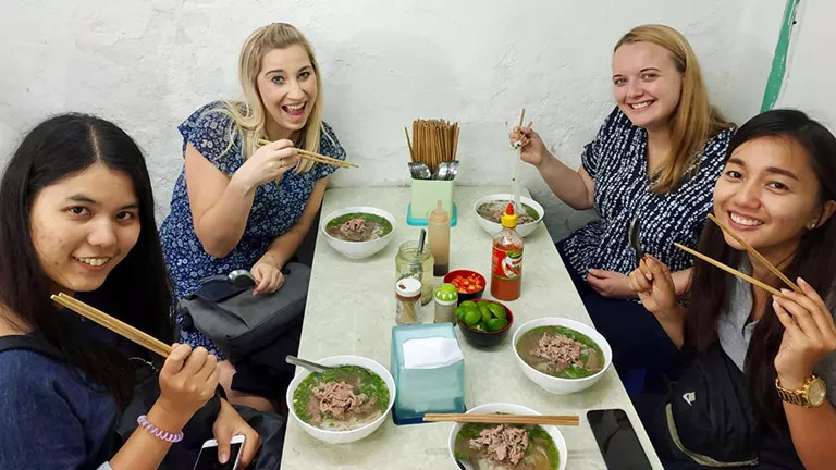 Four people hold chopsticks out in front of their bowls of pho on a shared table.
