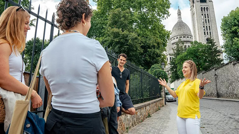Topdeck trip leader in a yellow uniform talks to a group of young people on a street in Paris, gesturing with their hands.