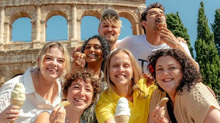 People posing together holding gelato and canolis in front of the Roman colosseum.