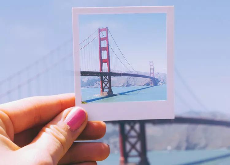 Topdeck traveler holding up a polaroid photograph of the Golden Gate Bridge with the real Golden Gate Bridge in the background.