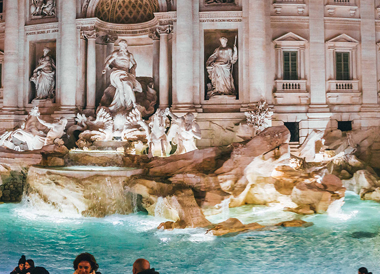 The famous Trevi Fountain of Rome lit up at night with a crowd of people surrounding it.