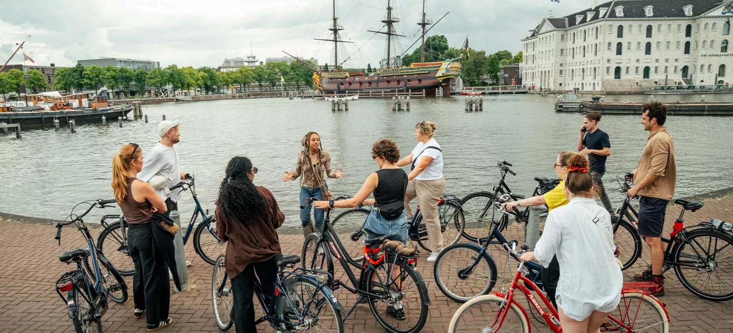Group of people standing with bikes, listening to person speak by the river in Amsterdam.