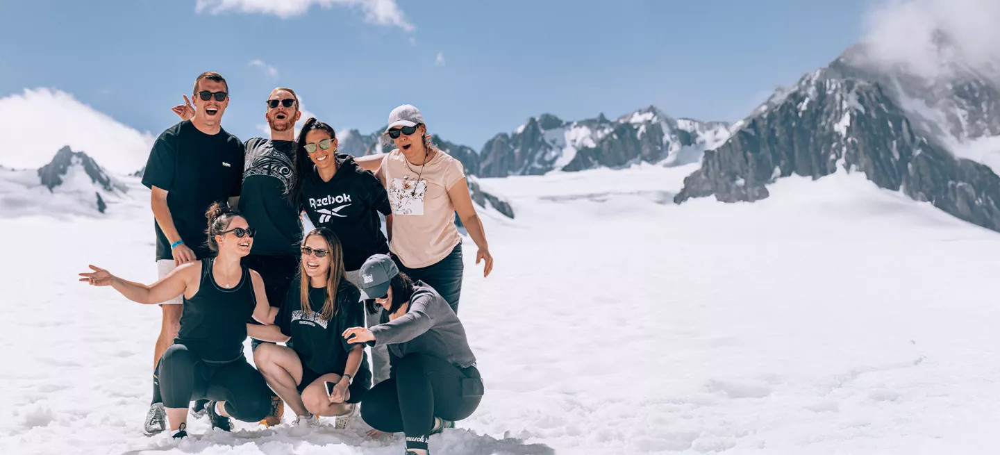 Group of seven people posing together smiling while standing on a the snow of a glacier in New Zealand.