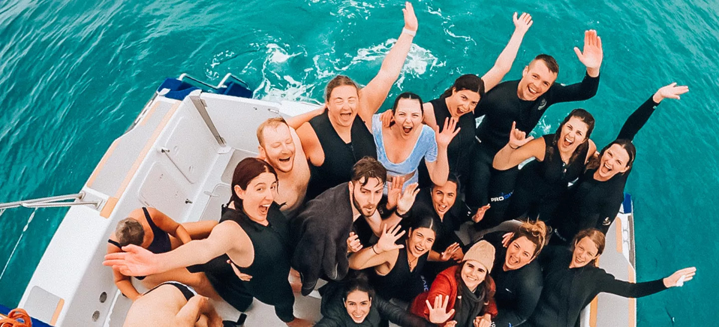 Group of people standing on the back of a boat wearing wetsuits with their hands in the air like they’re cheering.