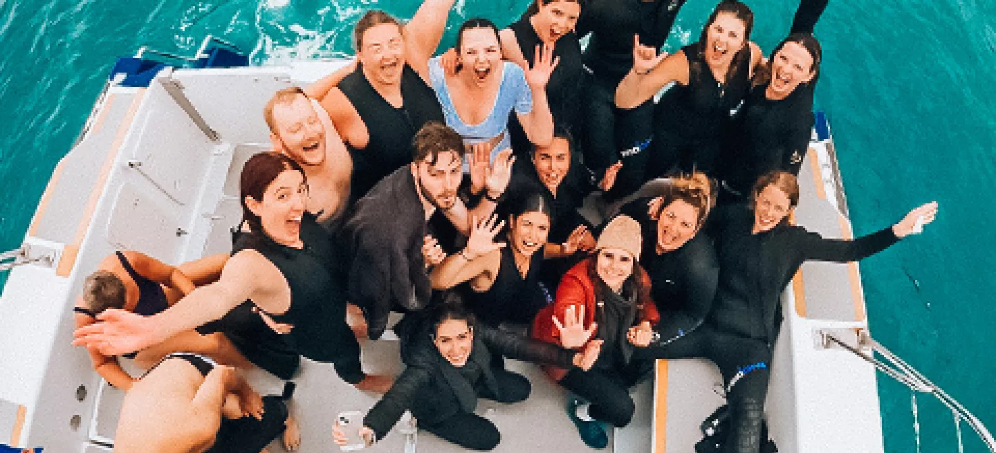 Group of people standing on the back of a boat wearing wetsuits with their hands in the air like they’re cheering.