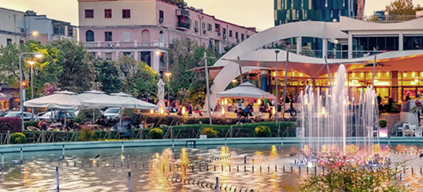 Pond with water feature in a public park with buildings in the background.