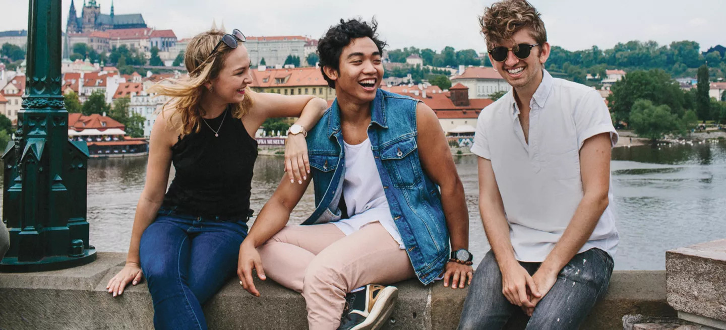 Three people sitting on a bridge in Prague with the river and a castle behind them.