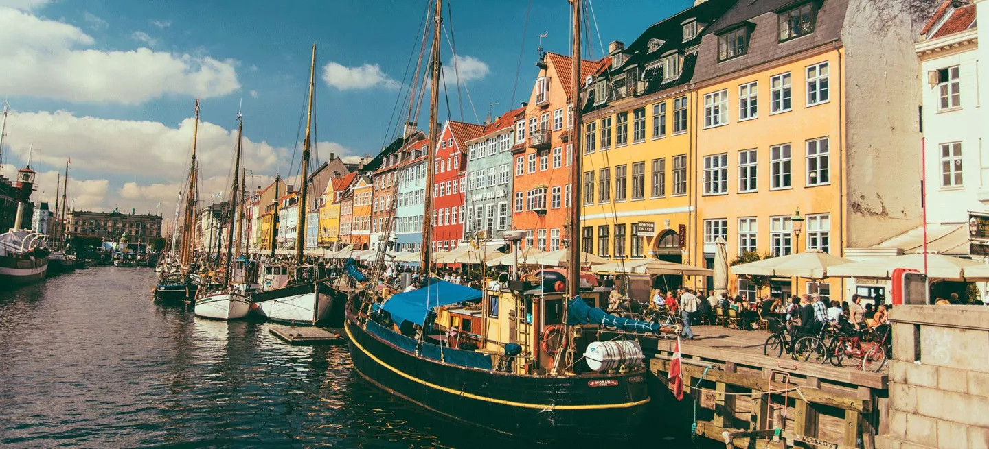 Boat with wooden mast docked in waterfront canal next to brightly coloured buildings in Copenhagen.