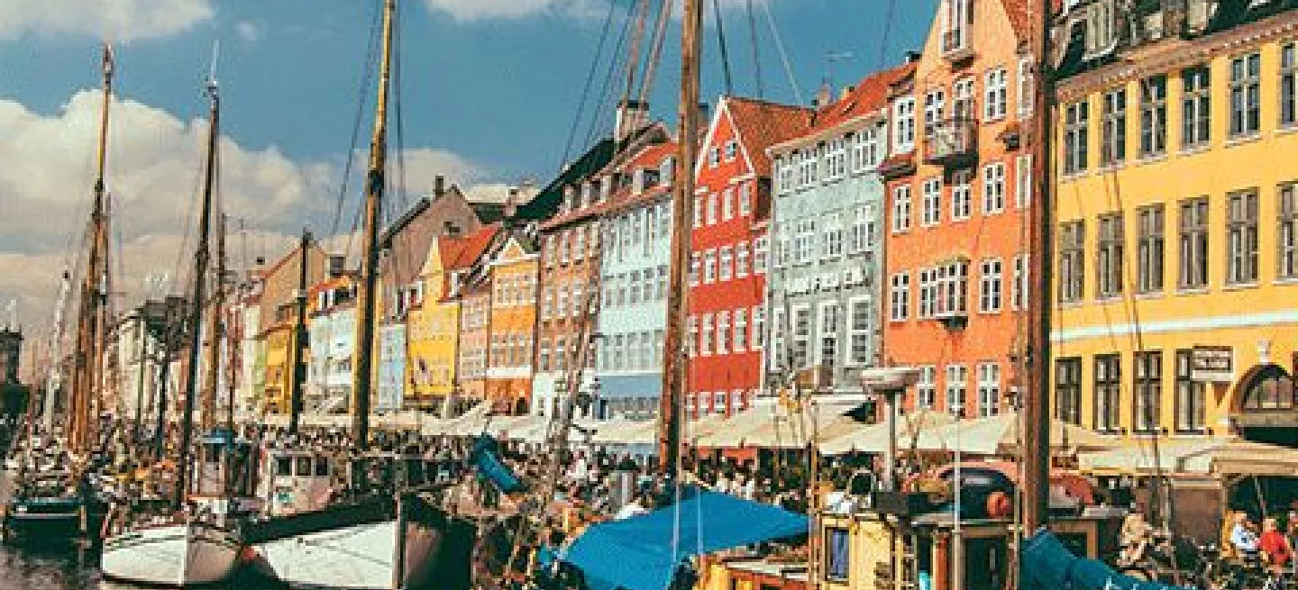 Boat with wooden mast docked in waterfront canal next to brightly coloured buildings in Copenhagen.