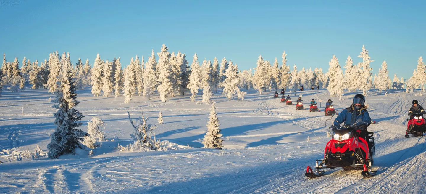 Single file line of people riding red skimobiles in the snow with an icy landscape behind them.