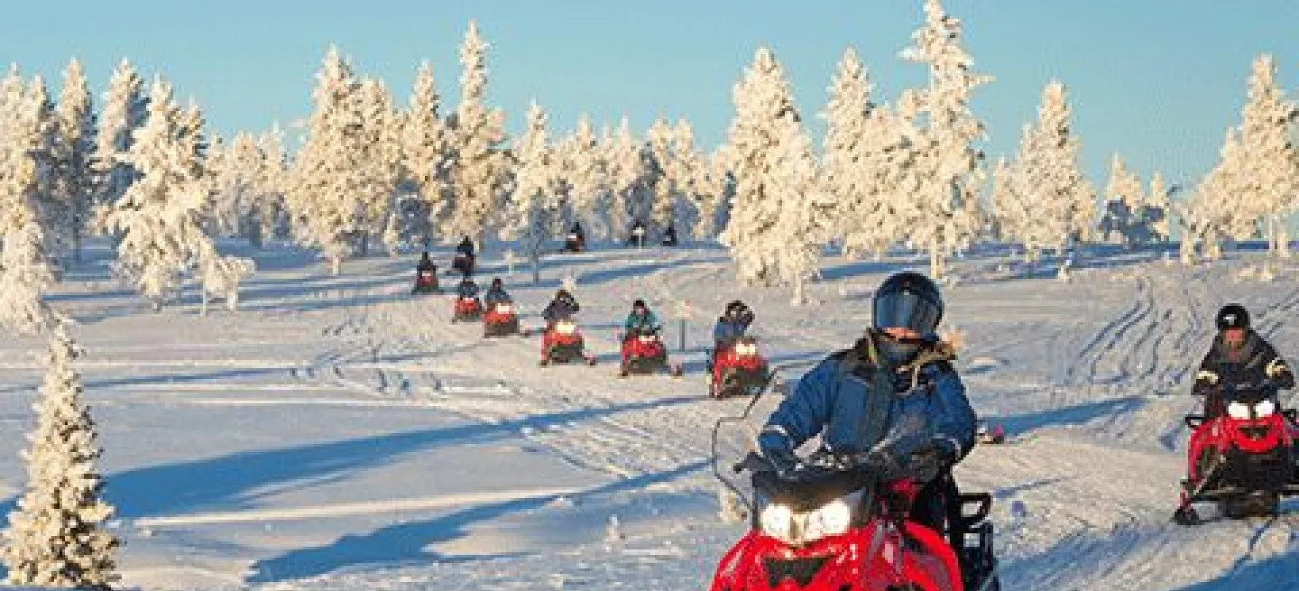 Single file line of people riding red skimobiles in the snow with an icy landscape behind them.