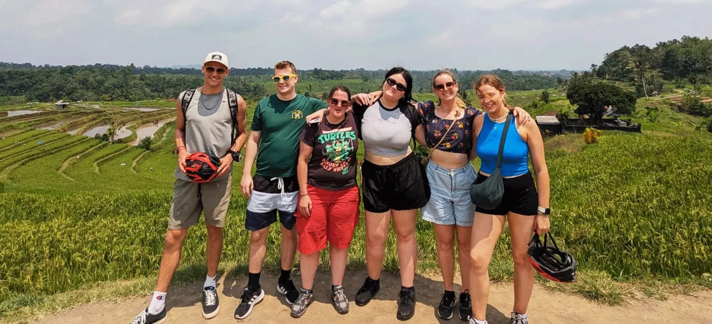 Group of people wearing active clothes pose together while holding bike helmets in front of a rice field in Bali, Indonesia.