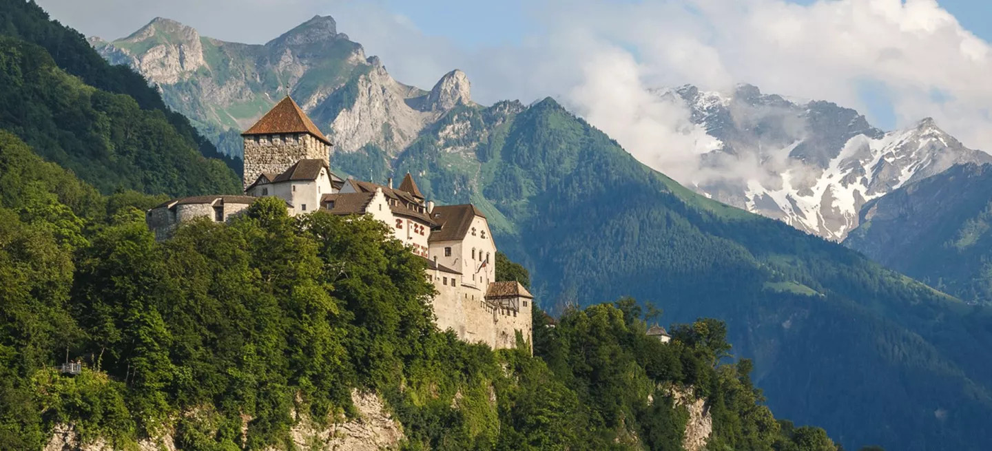 The Vaduz Castle on top of a hill surrounded by trees with mountains in the background.