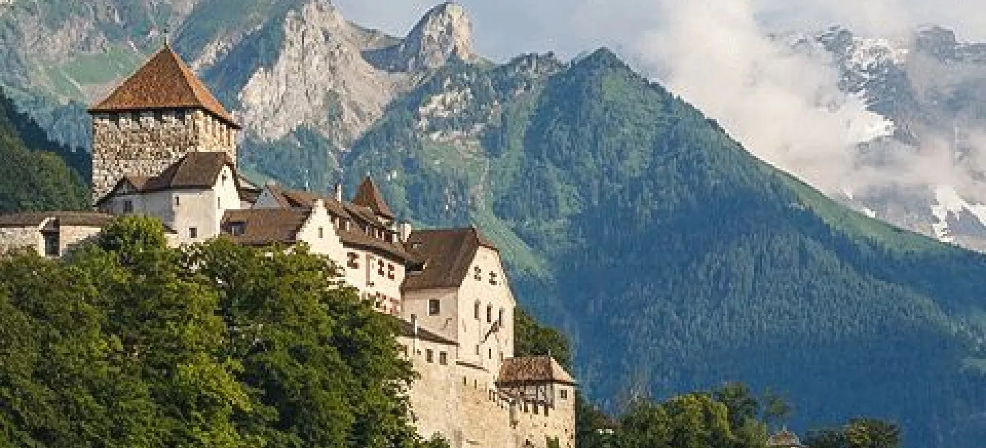 The Vaduz Castle on top of a hill surrounded by trees with mountains in the background.