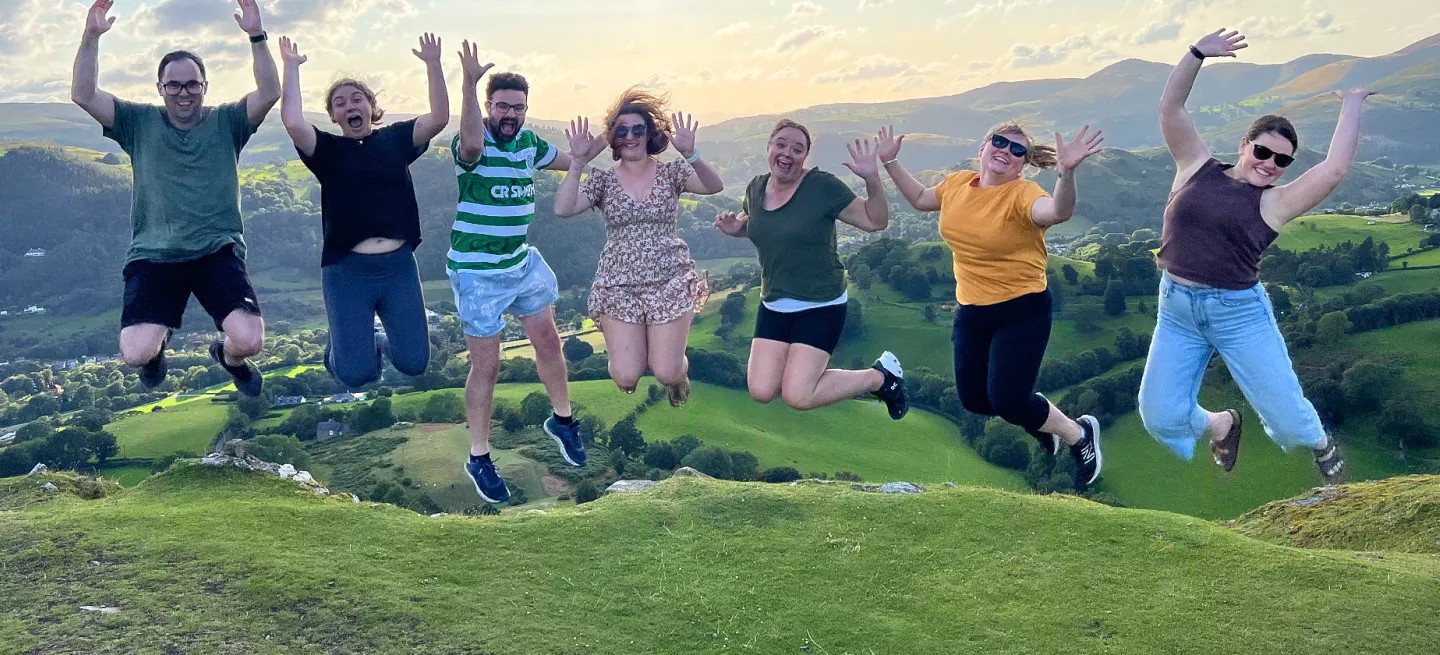 People jump in the air in a line with rolling hills behind them.