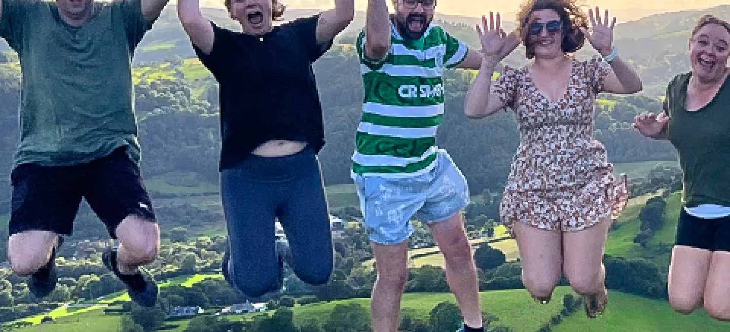 People jump in the air in a line with rolling hills behind them.