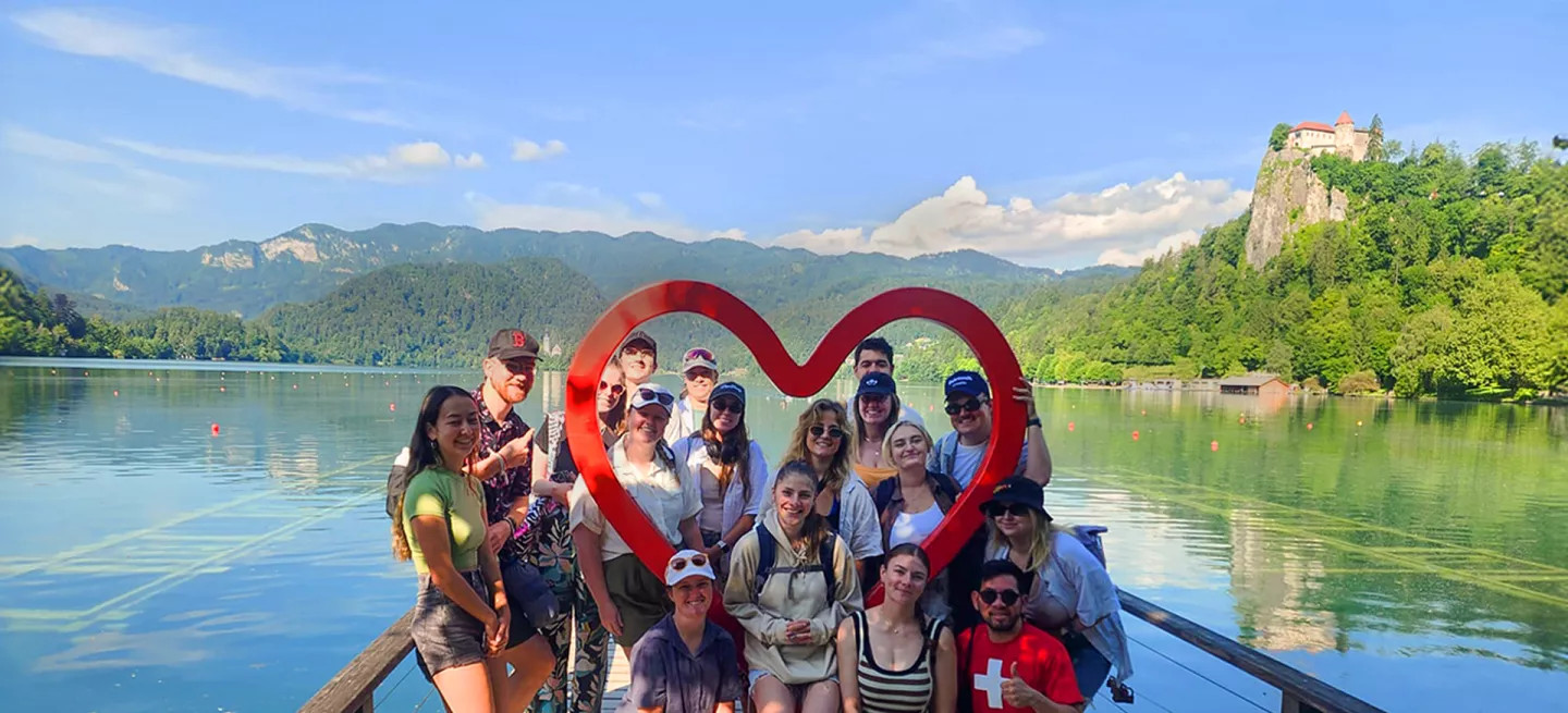 Group of people posing together around a red, wooden outline of a heart on the small wharf of a lake.