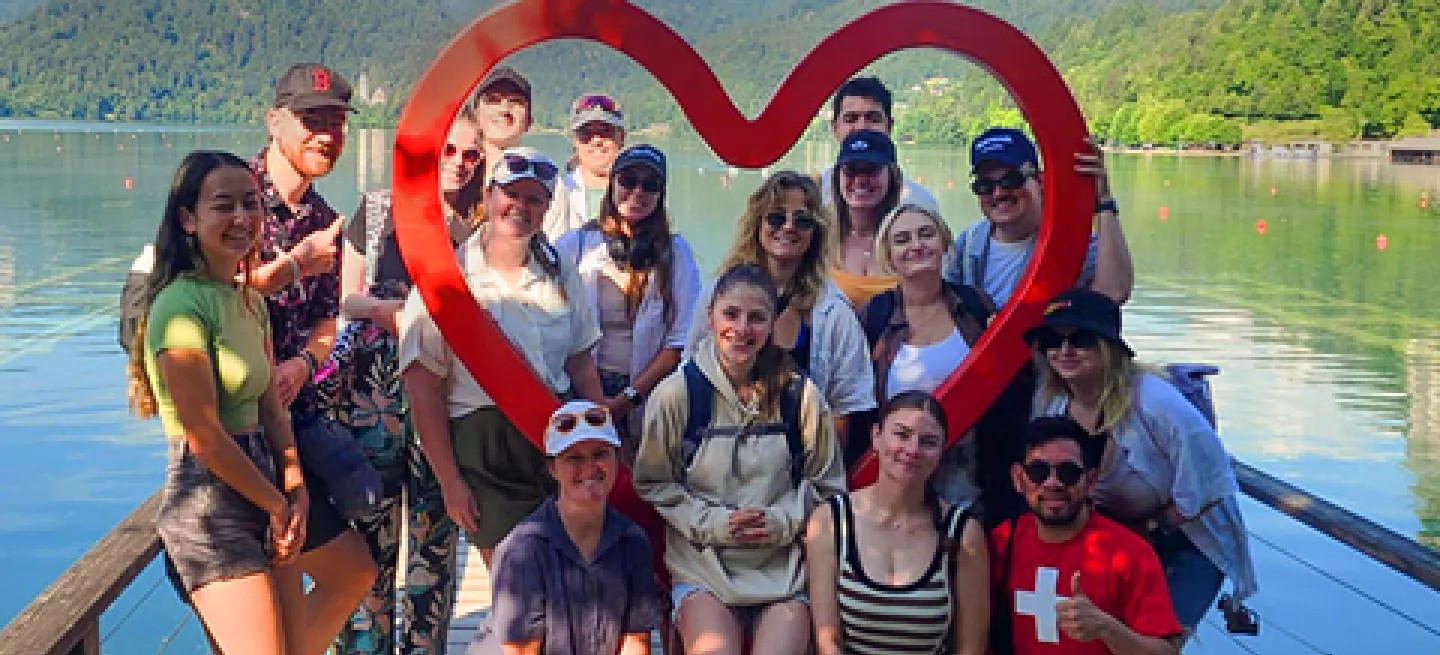 Group of people posing together around a red, wooden outline of a heart on the small wharf of a lake.