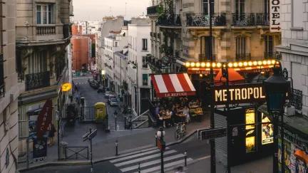 A wide view of a typical city street in Paris, with a busy restaurant and a sign for the Paris metro in view.