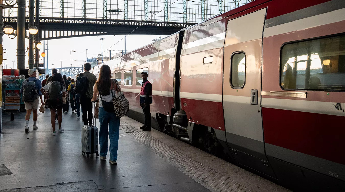 People walking with small bags along a platform with a red train to their right.