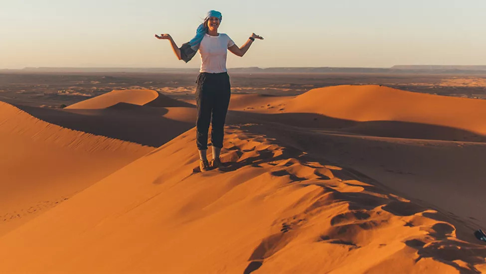 Woman standing smiling on sand dune ridge with her arms out and palms facing the sky.