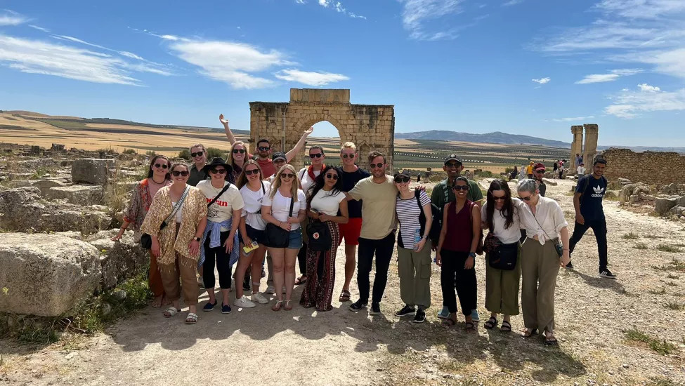 Group of people smiling with hands in the air, in front of the archaeological Site of Volubilis in Morocco.