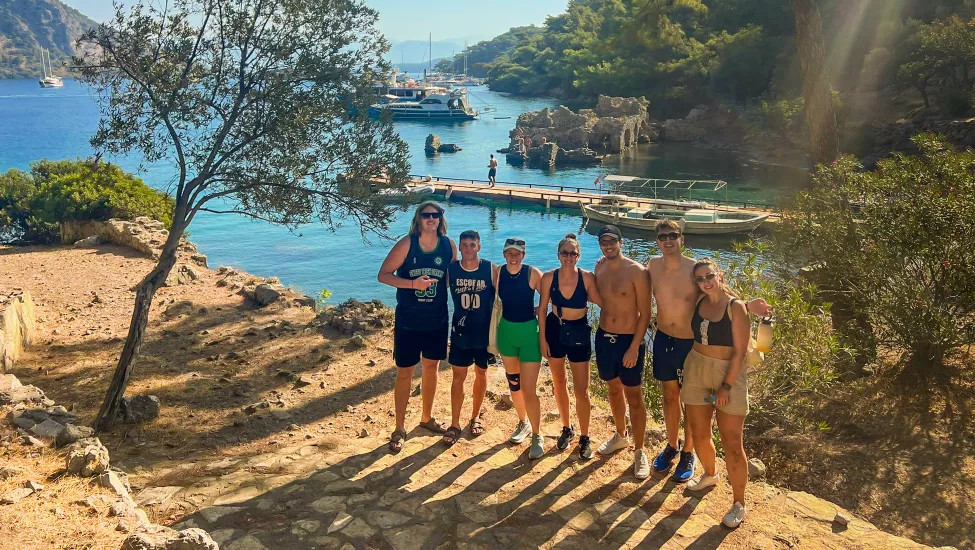 Seven people standing on a hill in the sun with the ocean and boat dock behind them.