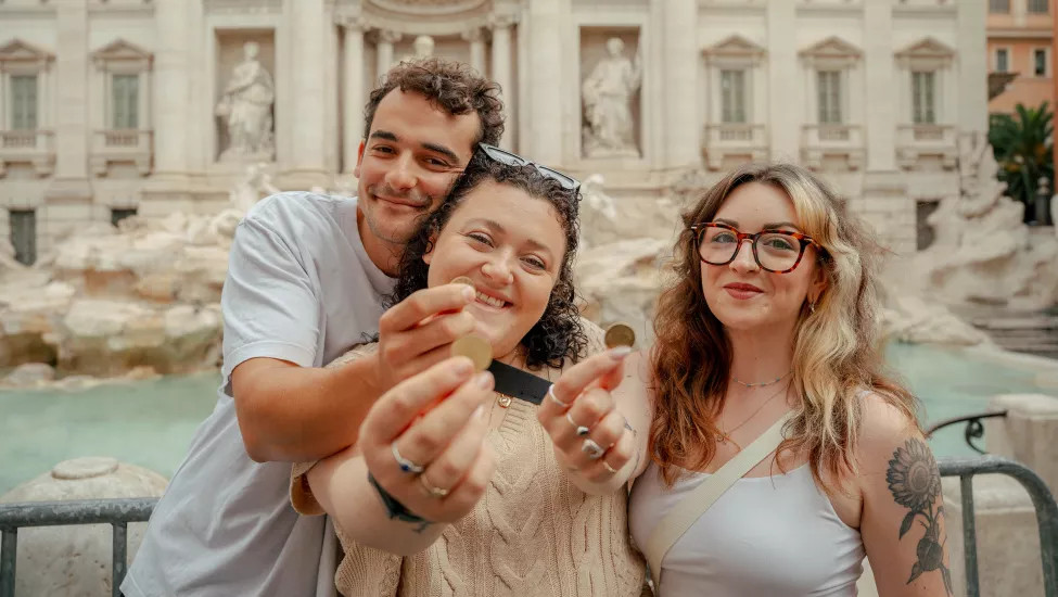 Three people smile while holding out coins in front of the Trevi Fountain.