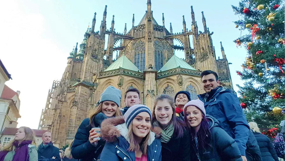 Group of people dressed in winter coats and smiling in front of St. Vitus Cathedral in Prague at Christmas time.