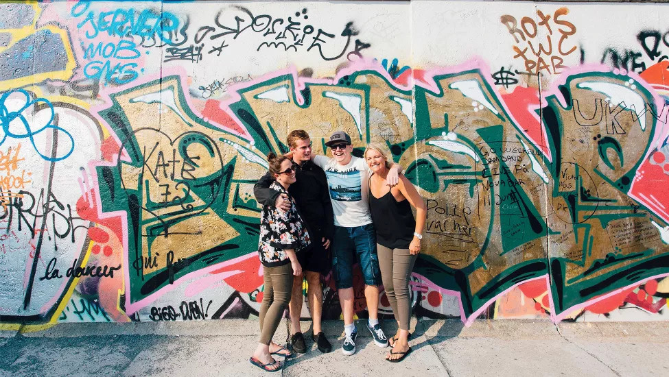 Four people stand together smiling in front of the graffitied Berlin Wall.