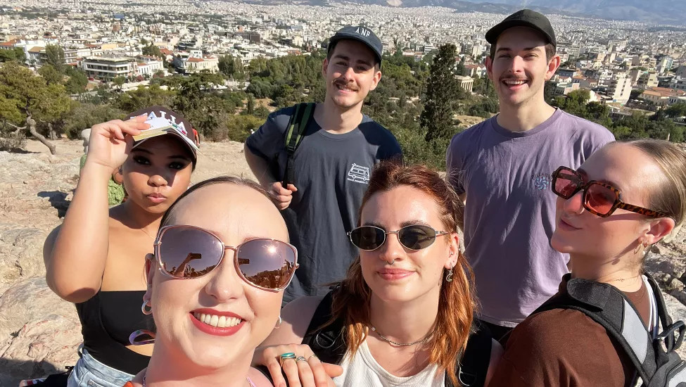 People smiling together for a photo on a hill by the Acropolis with a view over Athens behind them.