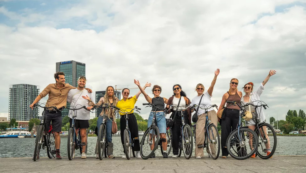 Group of people smiling with their hands in the air. They are sitting on bikes near the river in Amsterdam.