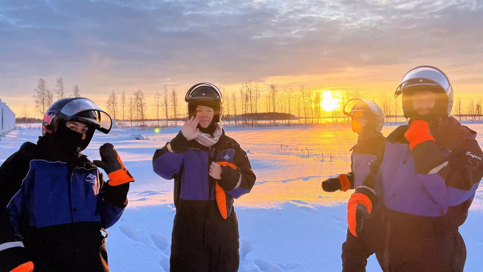 Four people posing in the snow in their snow suits with orange gloves and helmets.