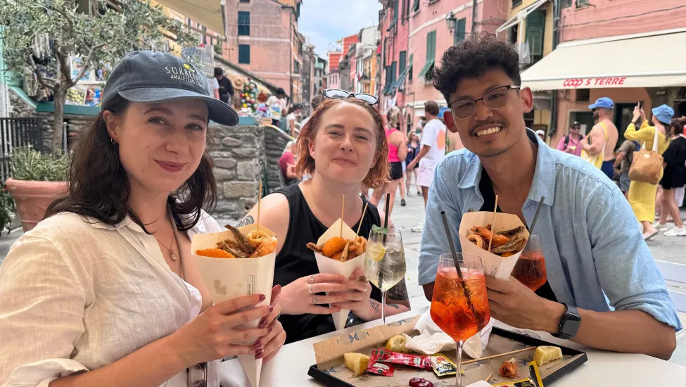 Three people smiling at a table in a Venice street. They hold fried street food and have spritz drinks in front of them.