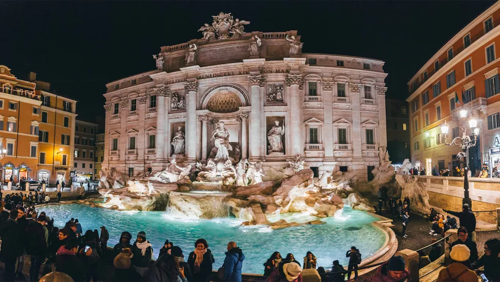 The famous Trevi Fountain of Rome lit up at night with a crowd of people surrounding it.