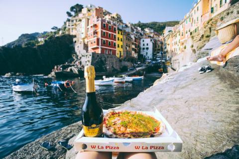 A person resting pizza and a bottle of prosecco on their legs with Italian houses and sea is in front of them