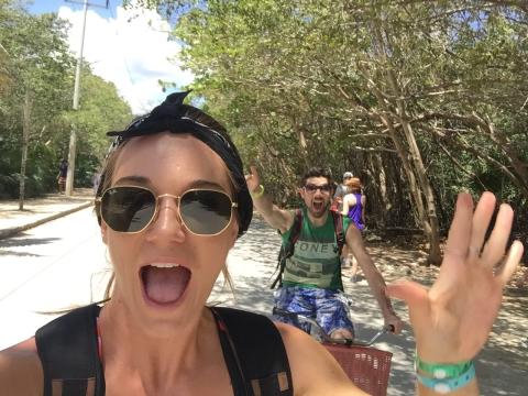 A woman smiling with her mouth open and wearing sunglasses. a man in the background is on a bike and raises a hand in the air