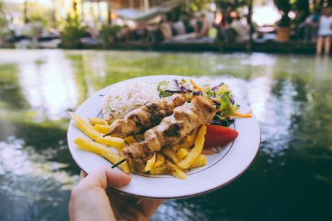 A hand holding a shish kebab with chips on a plate. the background is of a green river