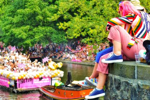 two people sit on the side of the river. In the background a boat is on the river with many people and balloons on it