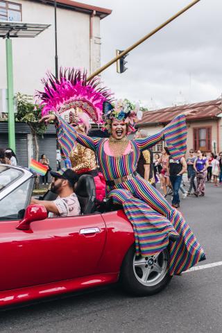 A drag queen sitting on the edge of a red car, wearing a striped rainbow jumpsuit