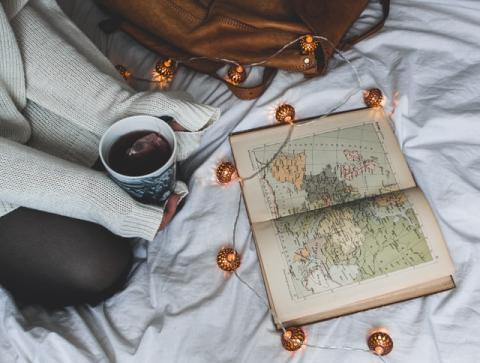 an open book showing a map on abed, a person sits in front of it, cupping a mug of tea