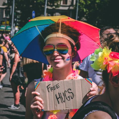 Woman wearing a rainbow umbrella hate, rainbow glasses and a sign that reads, 'Free Hugs'
