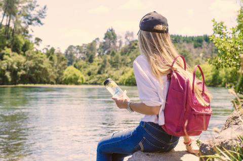 A girl sitting by the side of a lake, holding a water bottle and wearing a backpack