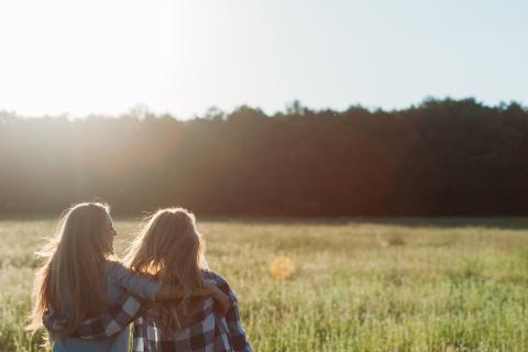 Two blonde woman embracing each other, with their backs to the camera, sitting in a field