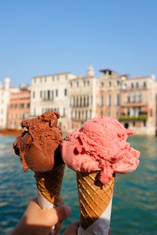 Two ice creams in cones (one chocolate, one strawberry) held up, with the ocean in the background
