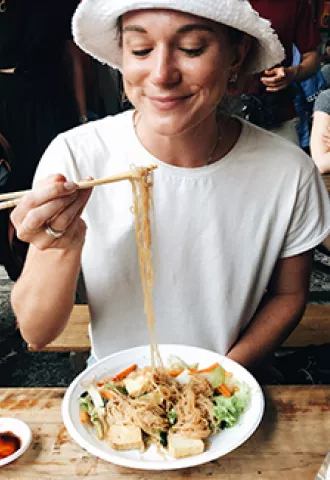 Person holds up noodles with a chopstick from a plate with vegetables and tofu.