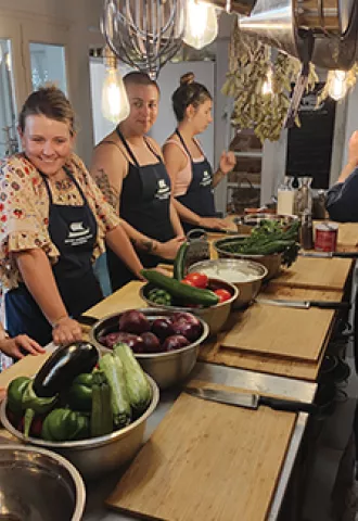 People standing on either side of a bench with aprons on at a cooking class. Fresh produce sits in bowls in the center.